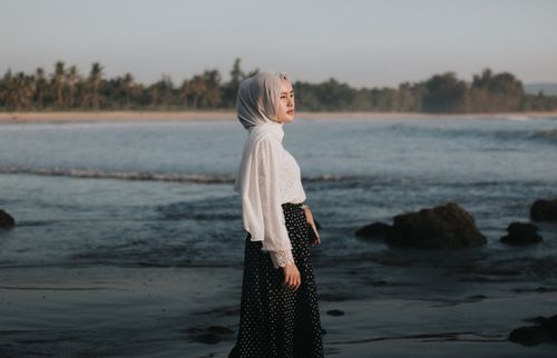 Free Woman in hijab standing on a tranquil beach, gazing at the sea under a clear sky. Stock Photo