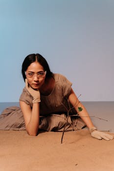 Artistic studio portrait of a woman in brown dress with gloves and goggles, posing with a tree branch on sand.
