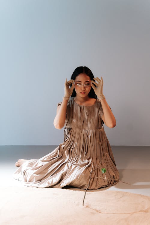 A Woman in Brown Dress Sitting Near the Sand while Holding Her Goggles