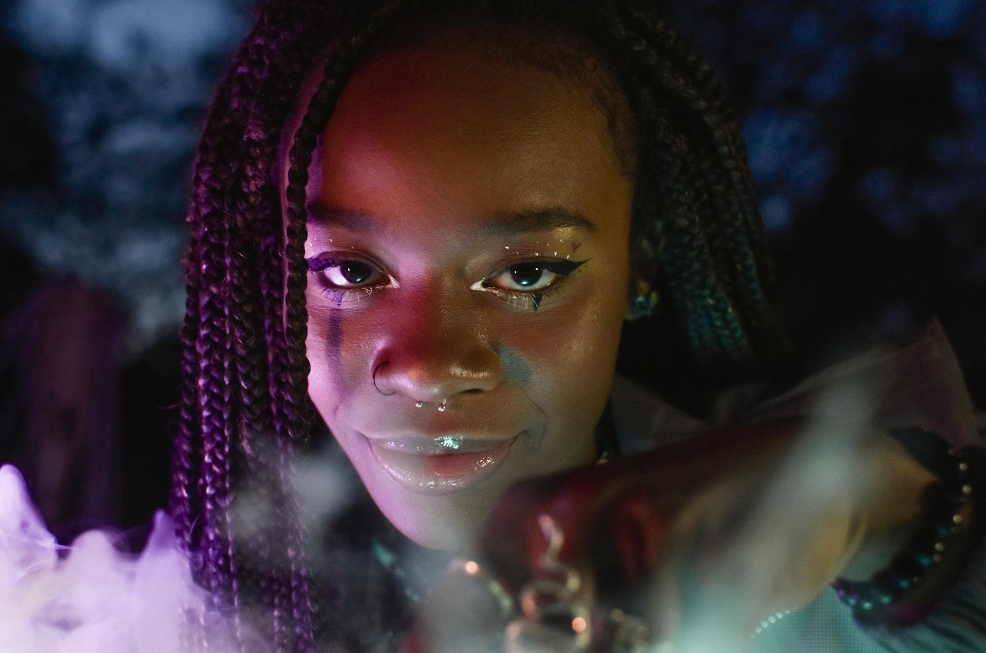 Close-up portrait of a smiling woman with braided hair and septum piercing, illuminated by vibrant lighting.