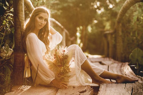 Free Side view of barefoot female in white dress sitting on footbridge with bouquet in forest and looking at camera Stock Photo