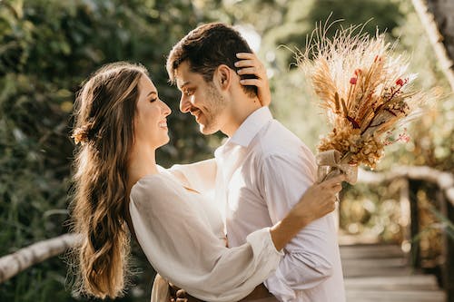 Side view of delightful newlyweds hugging and looking at each other while standing outside on blurred background