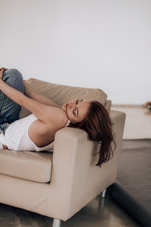 Side view of young female with dark brown hair in jeans and white shirt lying on leather beige sofa with eyes closed