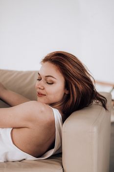 Side view of attractive calm woman with brown wavy hair and makeup wearing white tank top lying on comfortable leather sofa with closed eyes