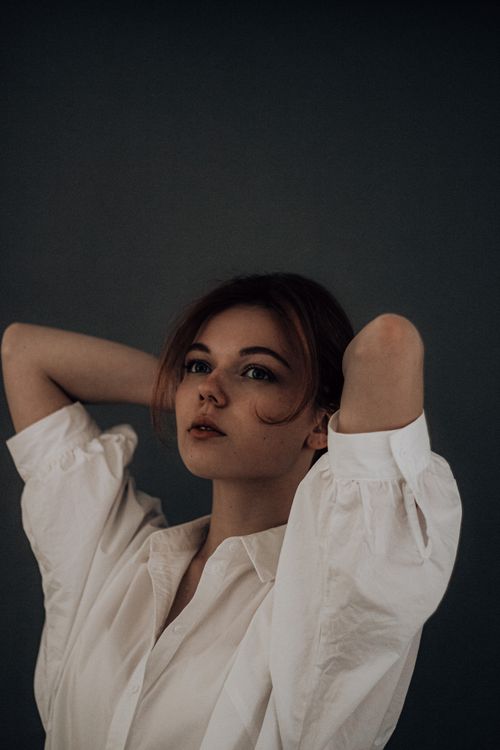 Free Attractive young female wearing white blouse with brown hair looking away with raised arms over head against gray wall Stock Photo