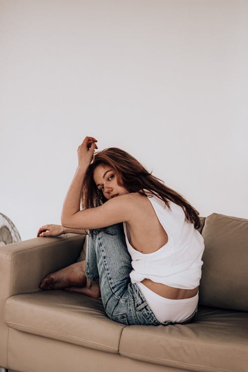 Side view of young female in casual clothes in white underwear and tank top sitting crossed legs on sofa with hands near head looking over shoulder at camera against white background