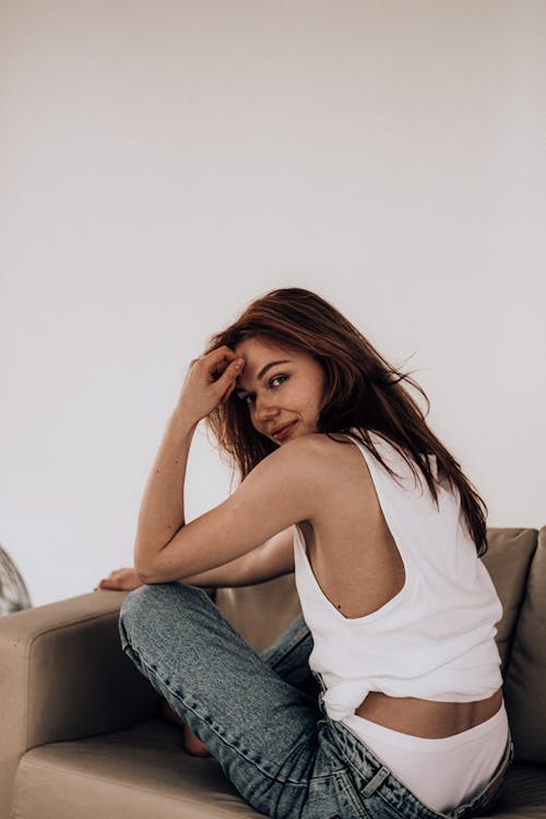 Free Side view of young woman in jeans and shirt sitting cross legged on comfortable couch and looking over shoulder at camera hand near forehead against white wall Stock Photo