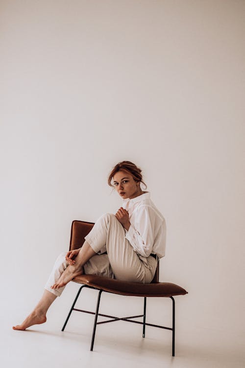Side view of young trendy female in white blouse and pants with brown hair pousing on chair against white background hand touching leg looking at camera