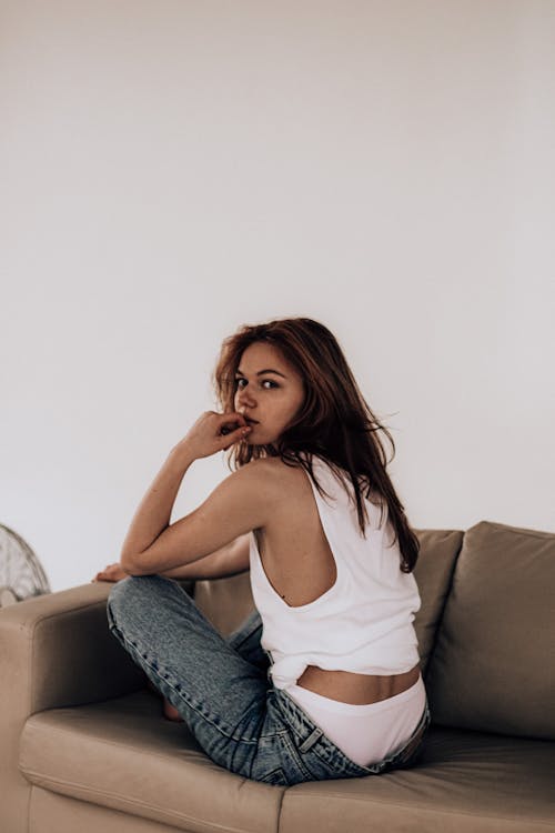Young thoughtful female with long dark hair in white t shirt and jeans sitting on couch with crossed legs and touching lips