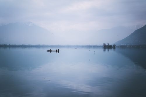 Free A serene lake scene with fishermen silhouetted at dawn, surrounded by misty mountains. Stock Photo