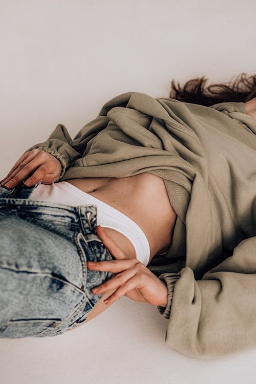 Crop unrecognizable female wearing casual hoodie with undressed belly taking off jeans while lying on white surface