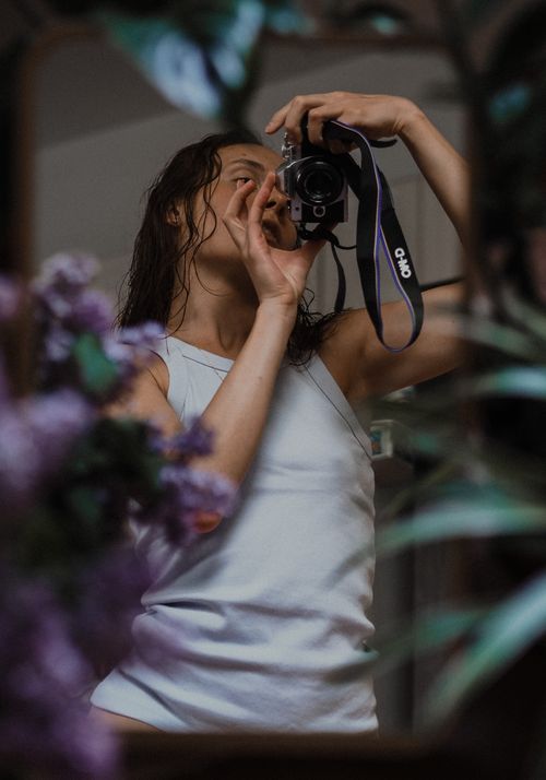 Free Young female photographer in casual white top with long dark hair taking photo on professional camera near window Stock Photo