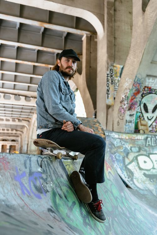 Free Young man in denim jacket sitting on skateboard in a graffiti-covered skate park. Stock Photo