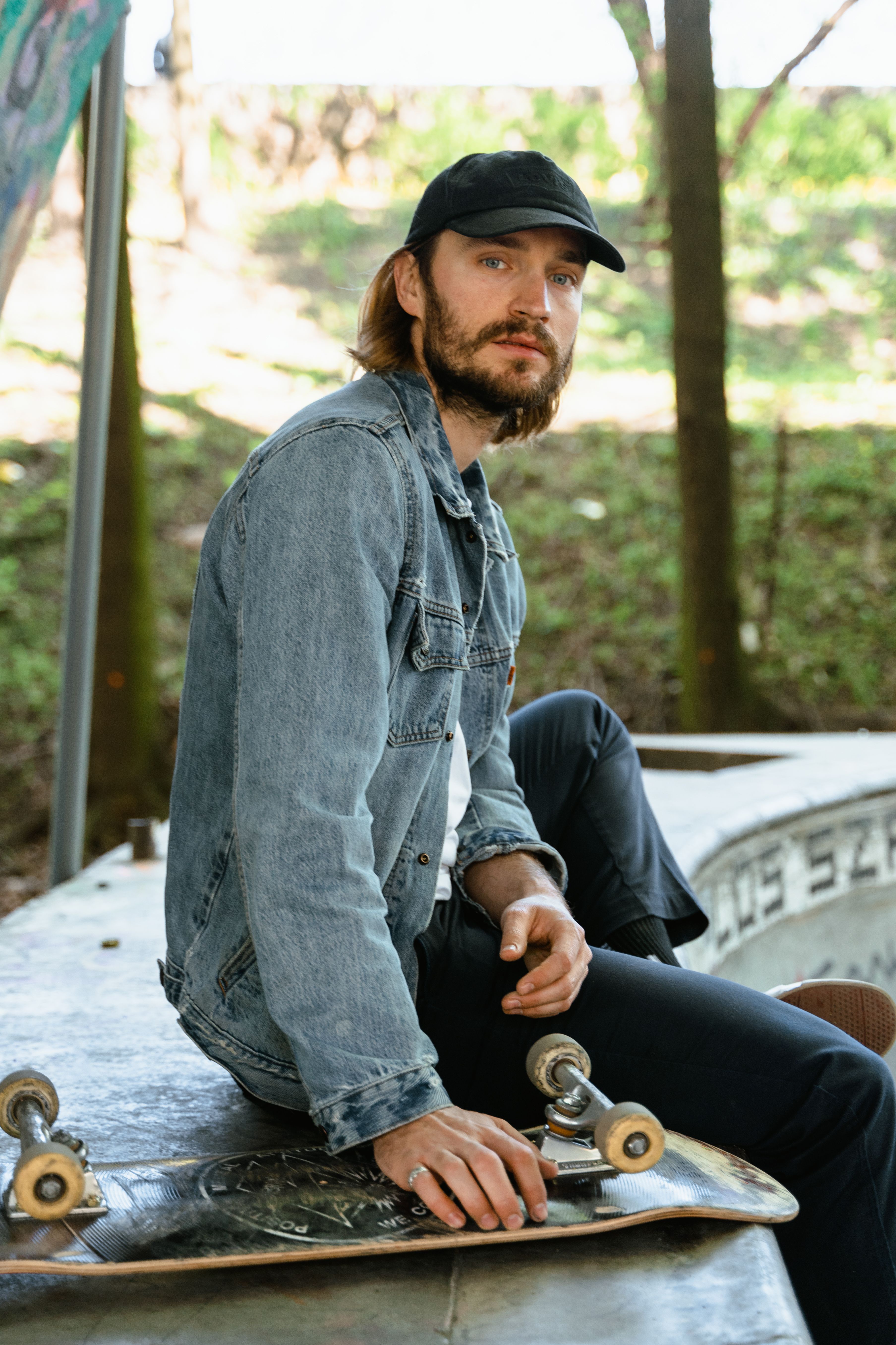 A man sitting casually with a skateboard, wearing a denim jacket and cap, outdoors.