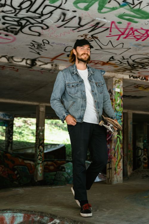 Free Young skateboarder posing in a graffiti-covered skate park, wearing a denim jacket and cap. Stock Photo