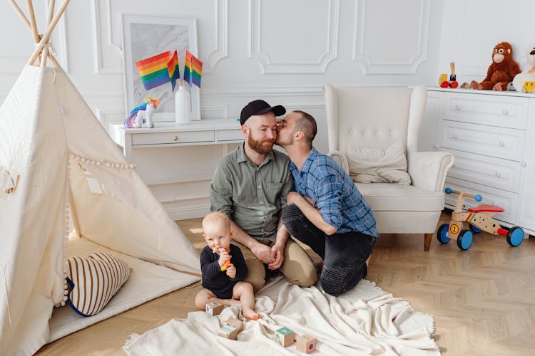 Man In Blue Plaid Shirt Kissing A Man In Green Dress Shirt Beside A Baby