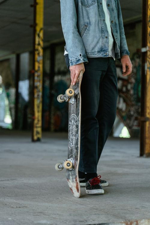 Free Person in denim jacket holding skateboard, standing in graffiti-filled urban area. Stock Photo