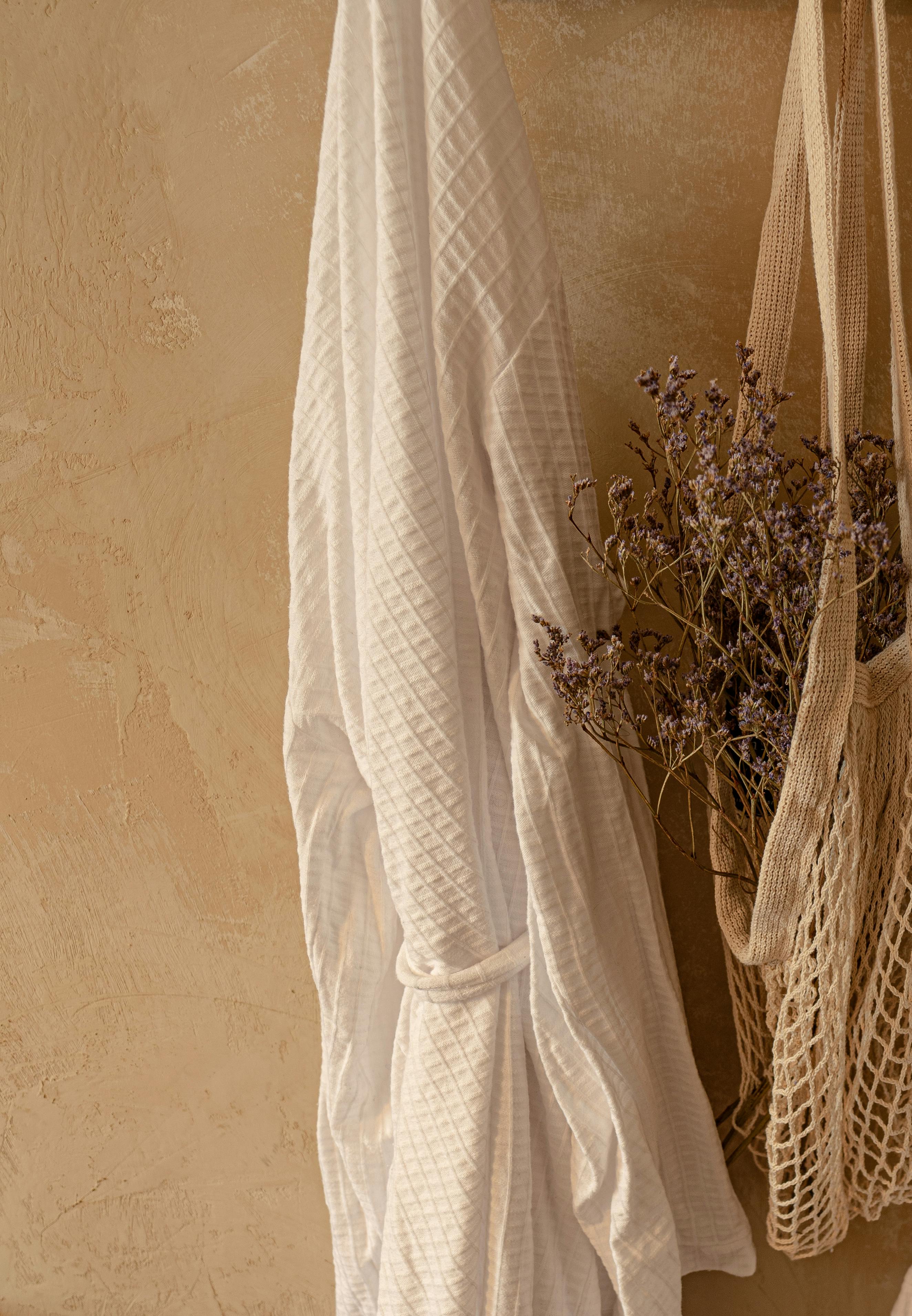Close-up of hanging dried flowers in a net bag with a white robe on a textured wall.