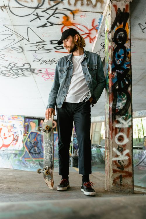 Free Young skater in denim jacket posing with skateboard in a graffiti-covered urban setting, embodying street style. Stock Photo