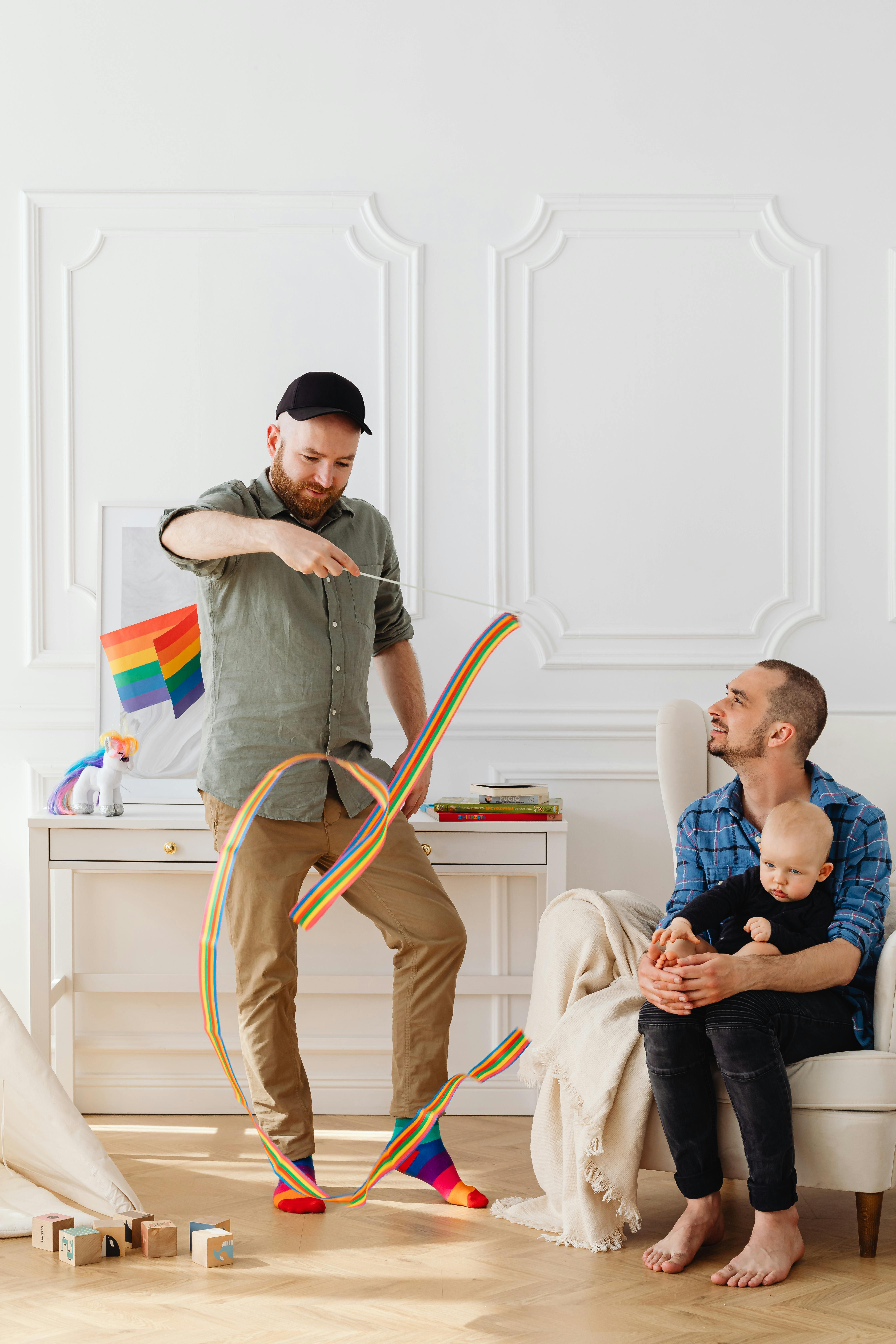A joyful gay couple playing with their child in a brightly lit room adorned with toys and a rainbow flag.
