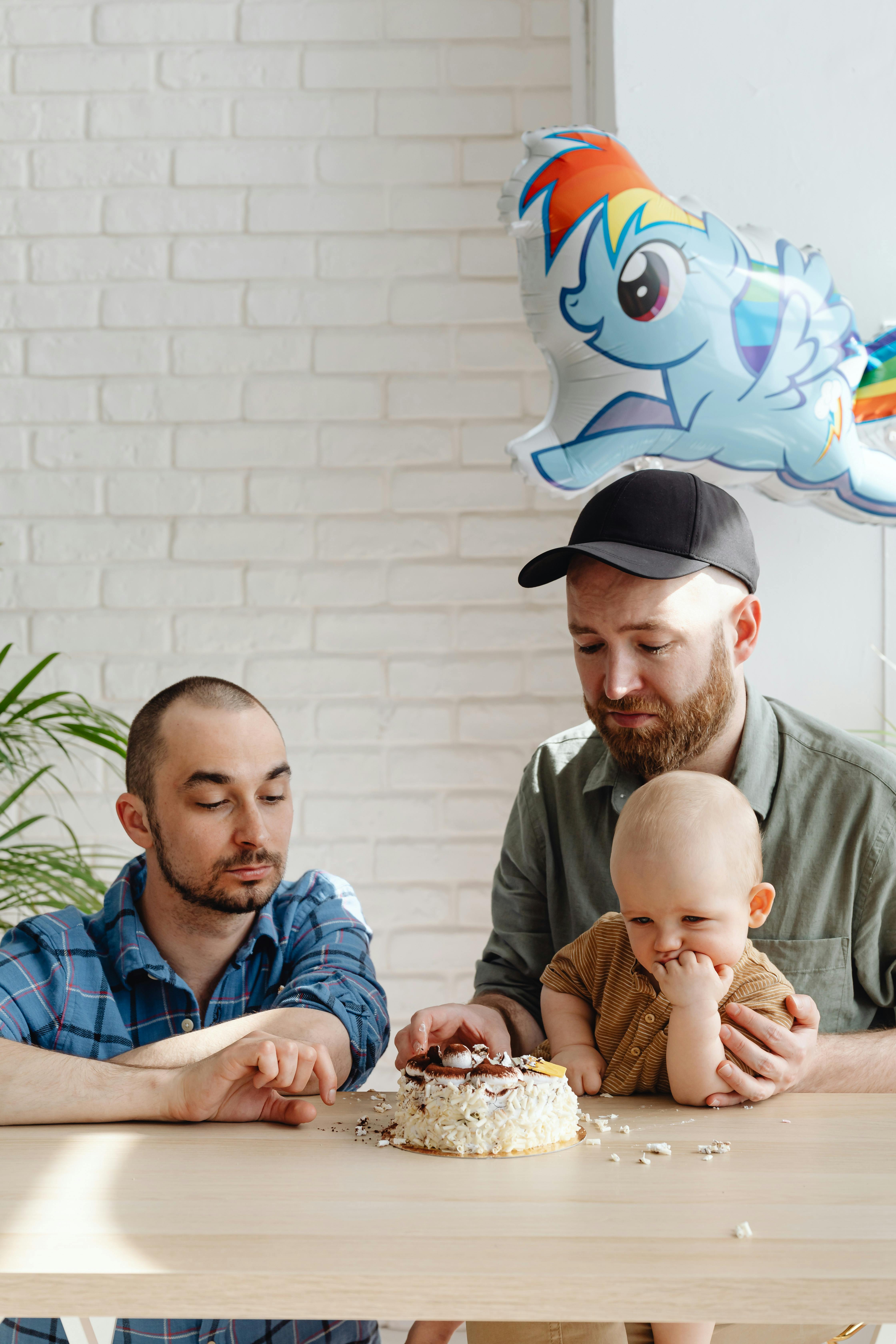 a same sex couple looking at the cake on the table with their son