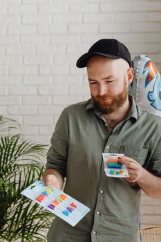 Bearded man in casual wear holding colorful candles indoor with plants.