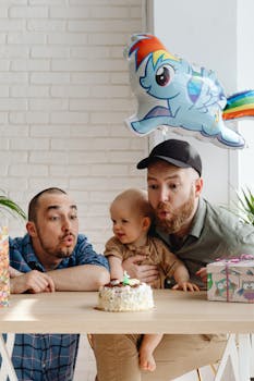 A joyful family moment capturing a birthday celebration with two men and a toddler blowing candles on a cake, adorned with a rainbow-themed balloon.