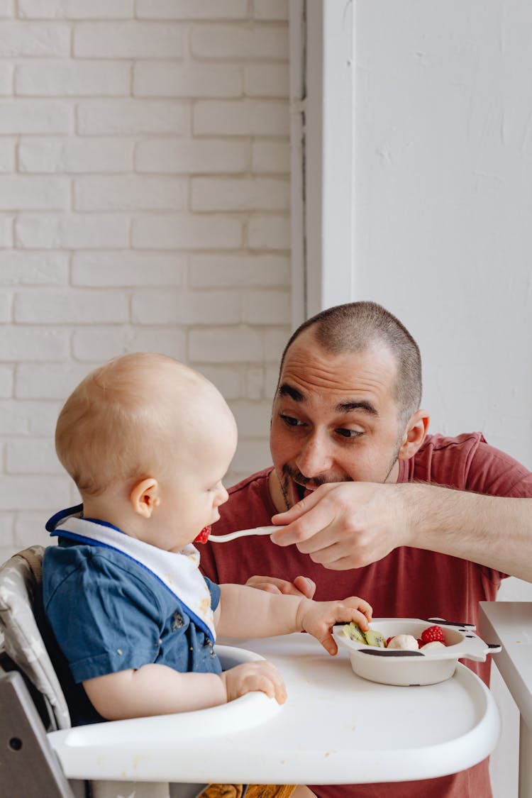 Man In Red Shirt Feeding Baby In Blue Shirt