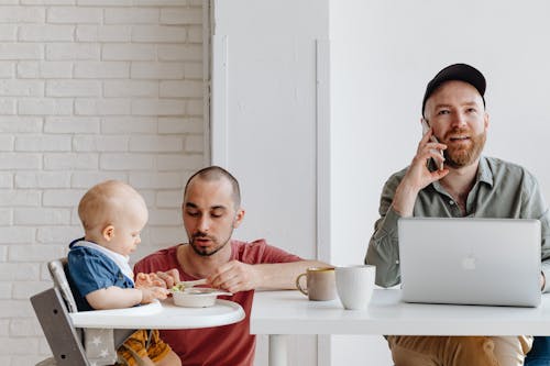 Father Feeding a Baby Next to His Partner Working on a Laptop