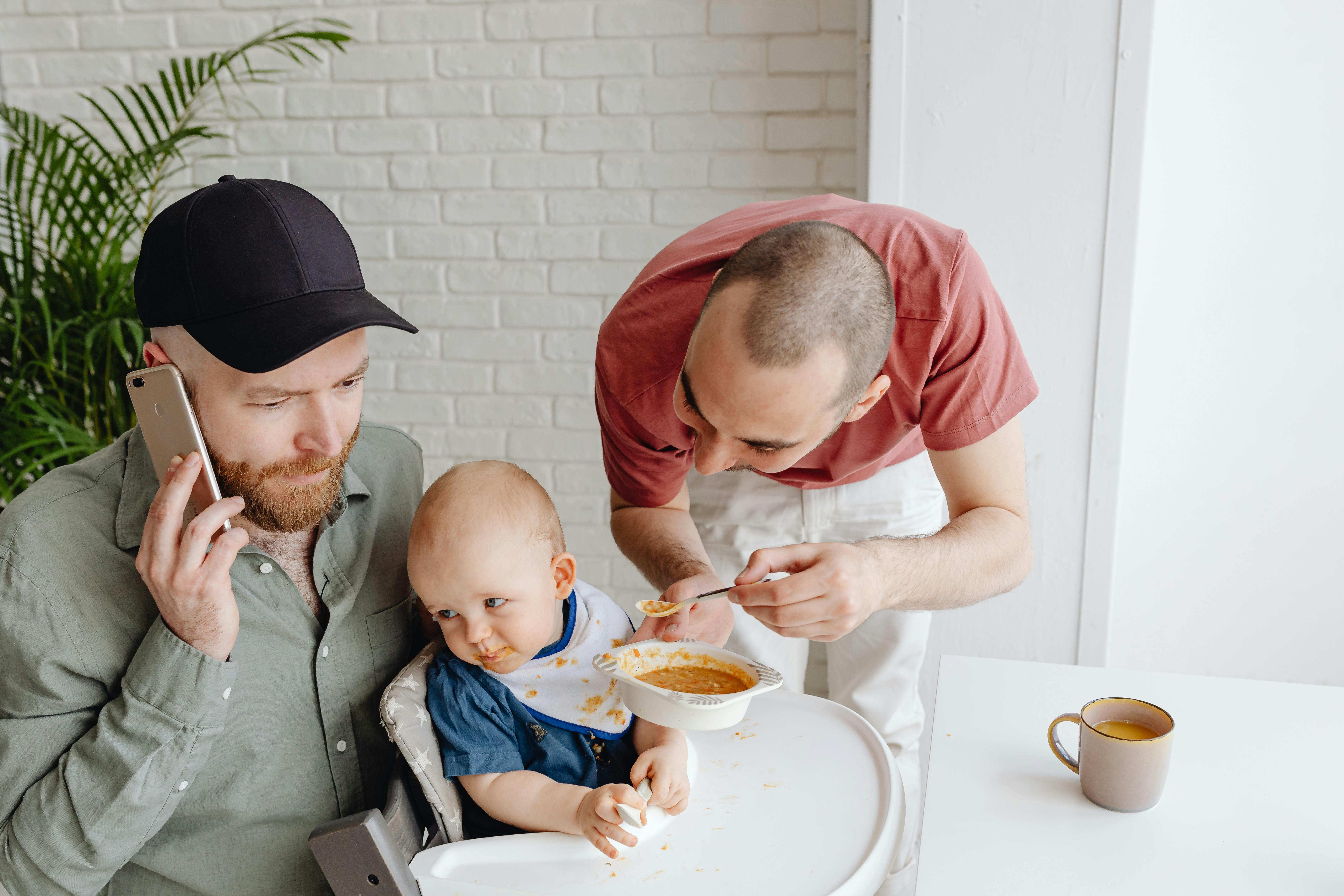 father talking on the phone next to a fussy baby spoon fed by his father