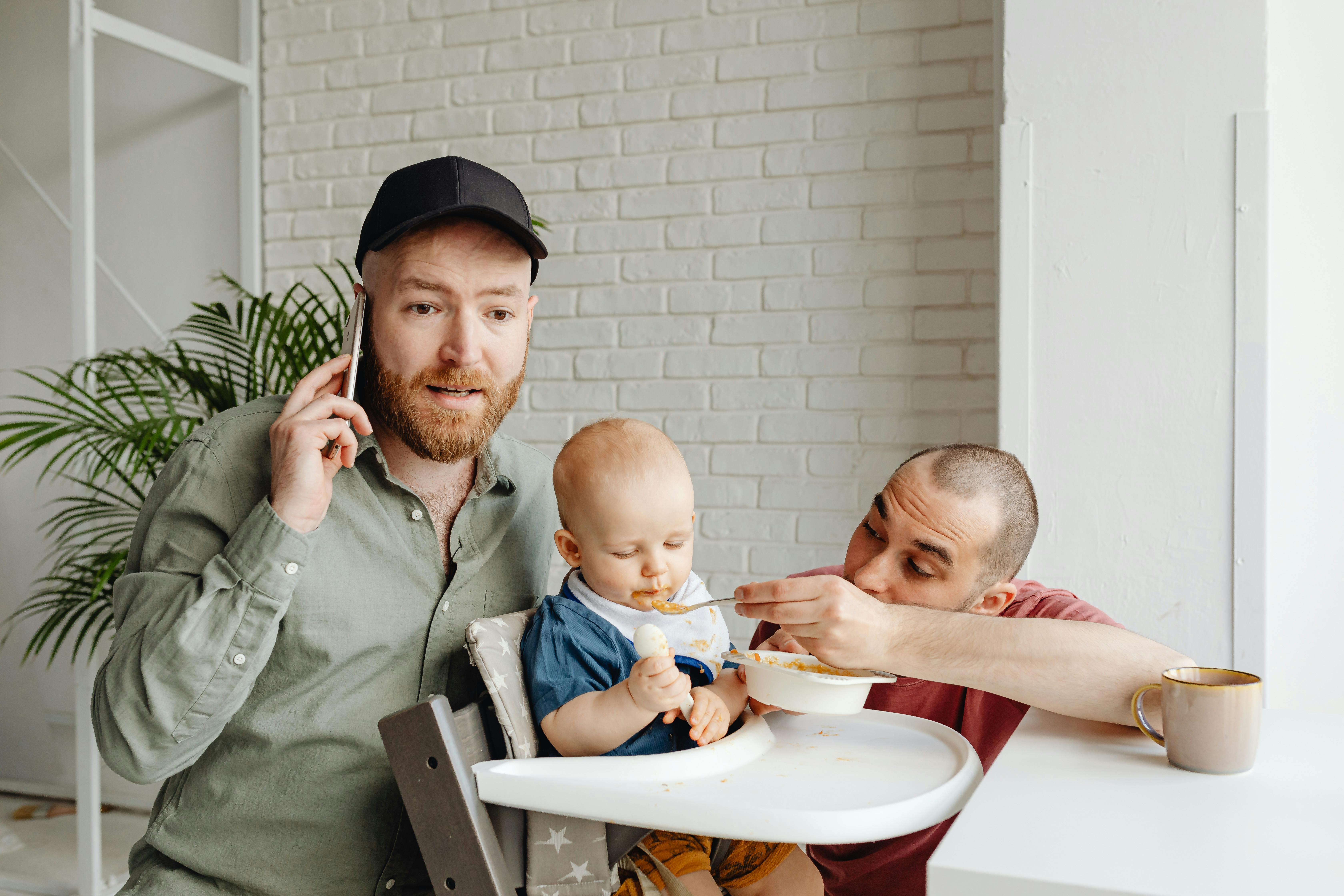 man in gray button up shirt talking on the cellphone beside baby in high chair