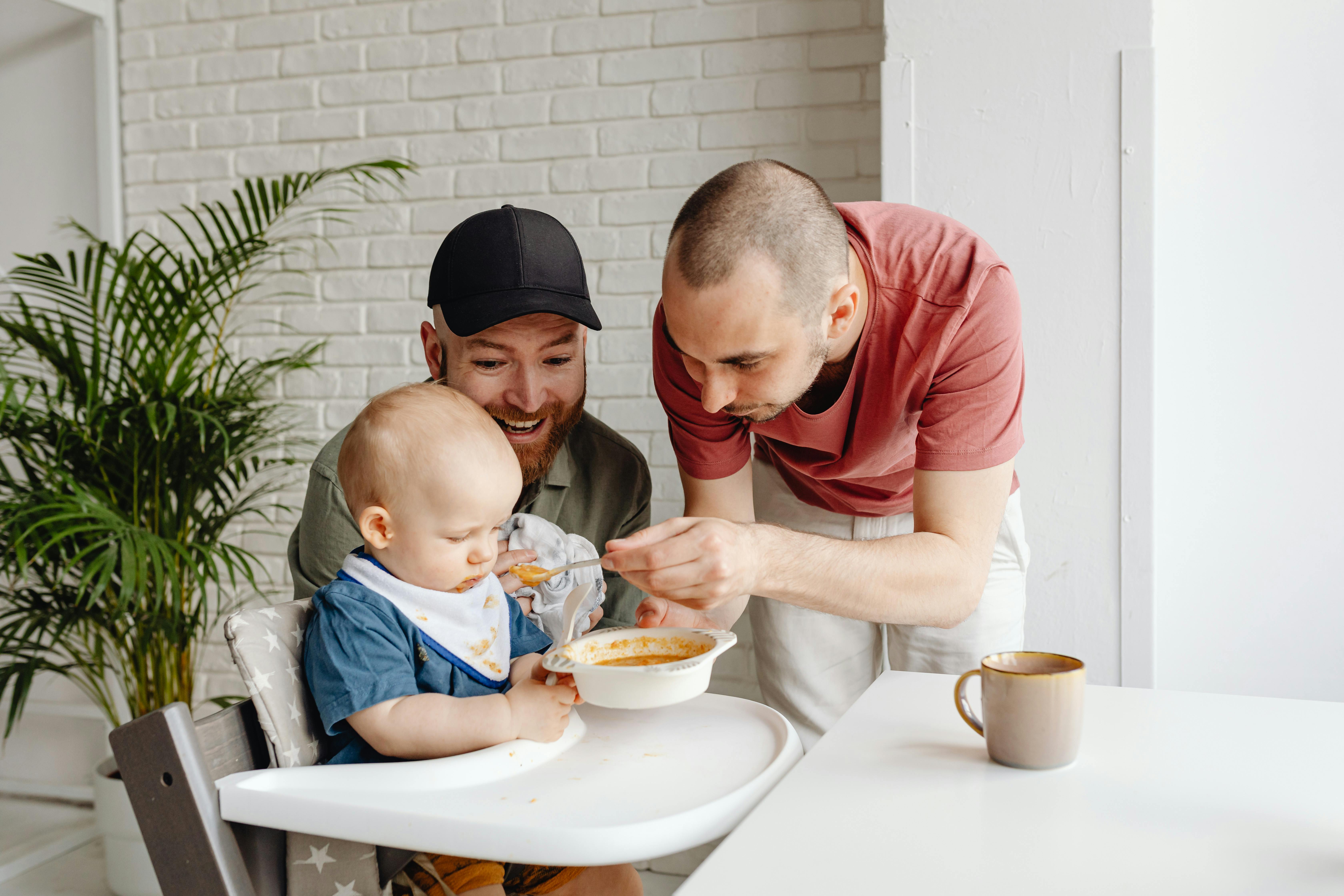 man in red crew neck t shirt feeding a baby beside man in green shirt