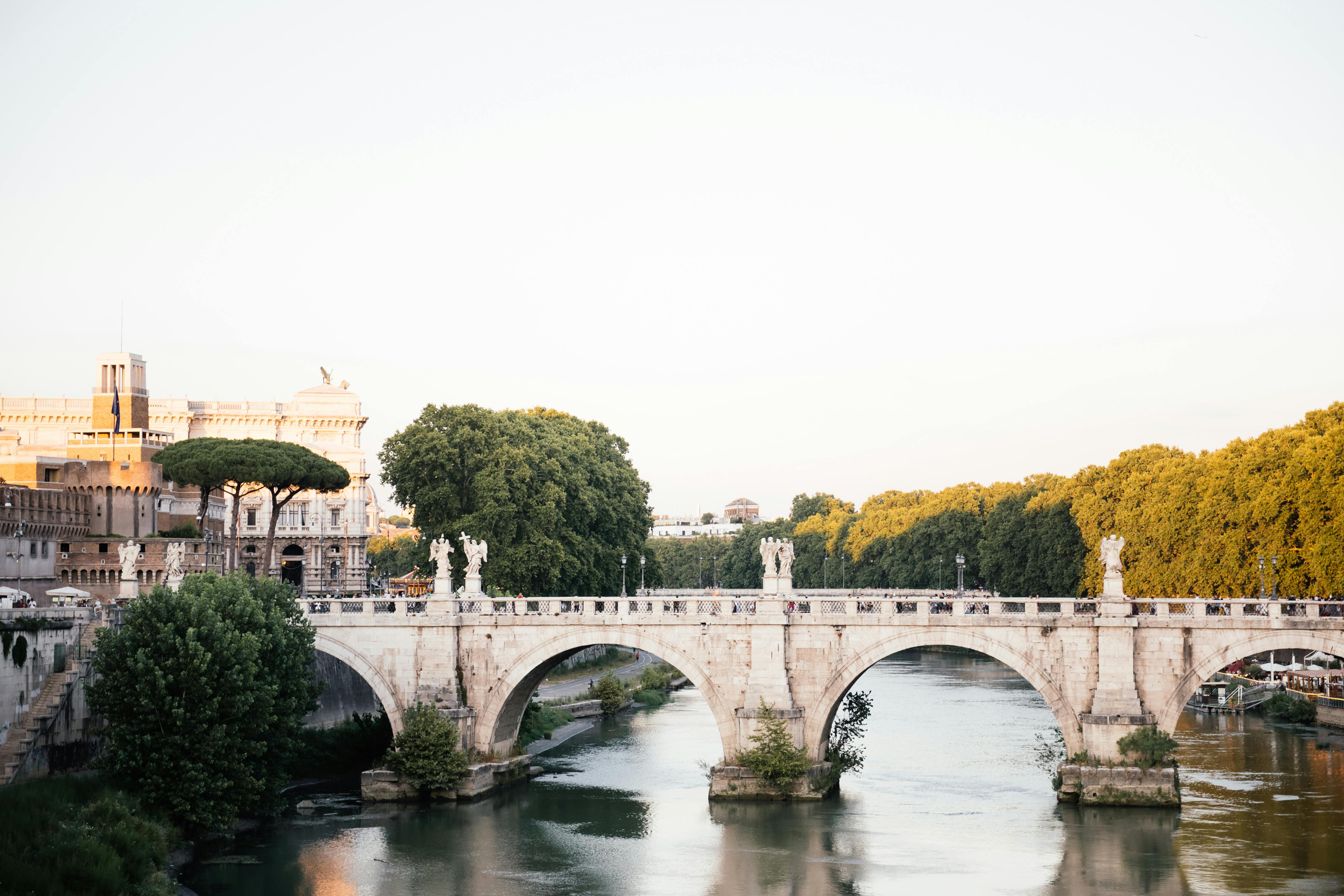 Picturesque view of the historic bridge over the Tiber River with Roman architecture and lush trees.