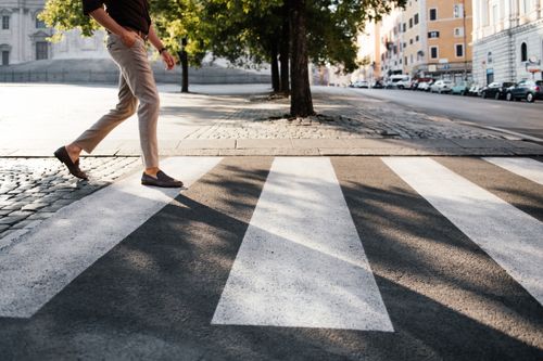 Free A person walking across a sunlit pedestrian crossing in a quiet city street. Stock Photo