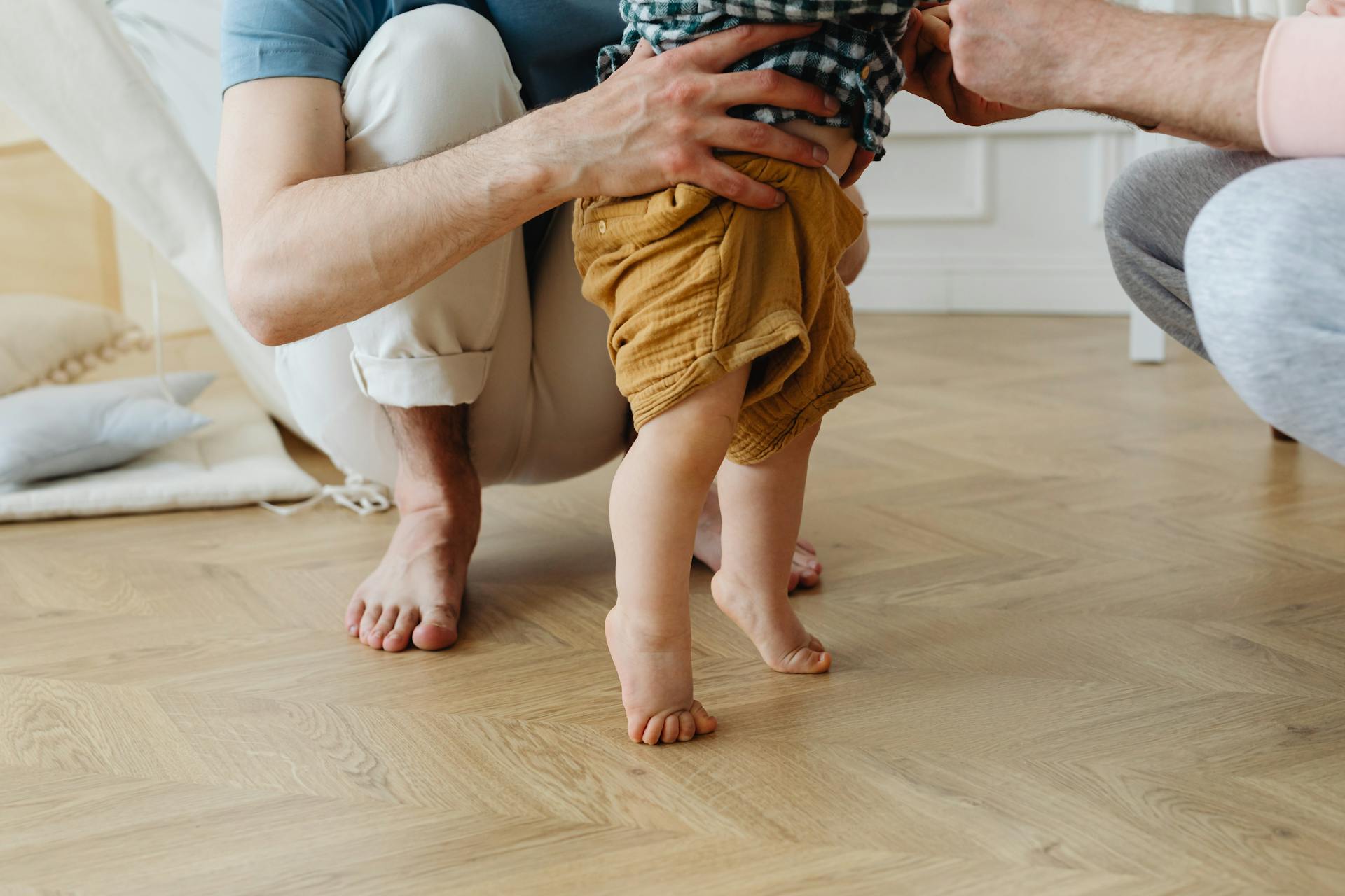 A heartwarming scene of a father helping his baby take first steps barefoot on a wooden floor.