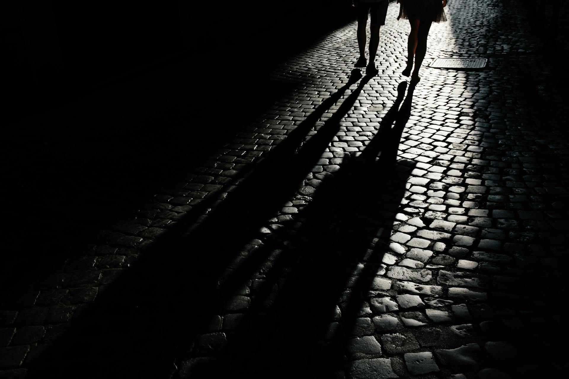 Two people walking on a cobblestone street casting long shadows in the darkness.