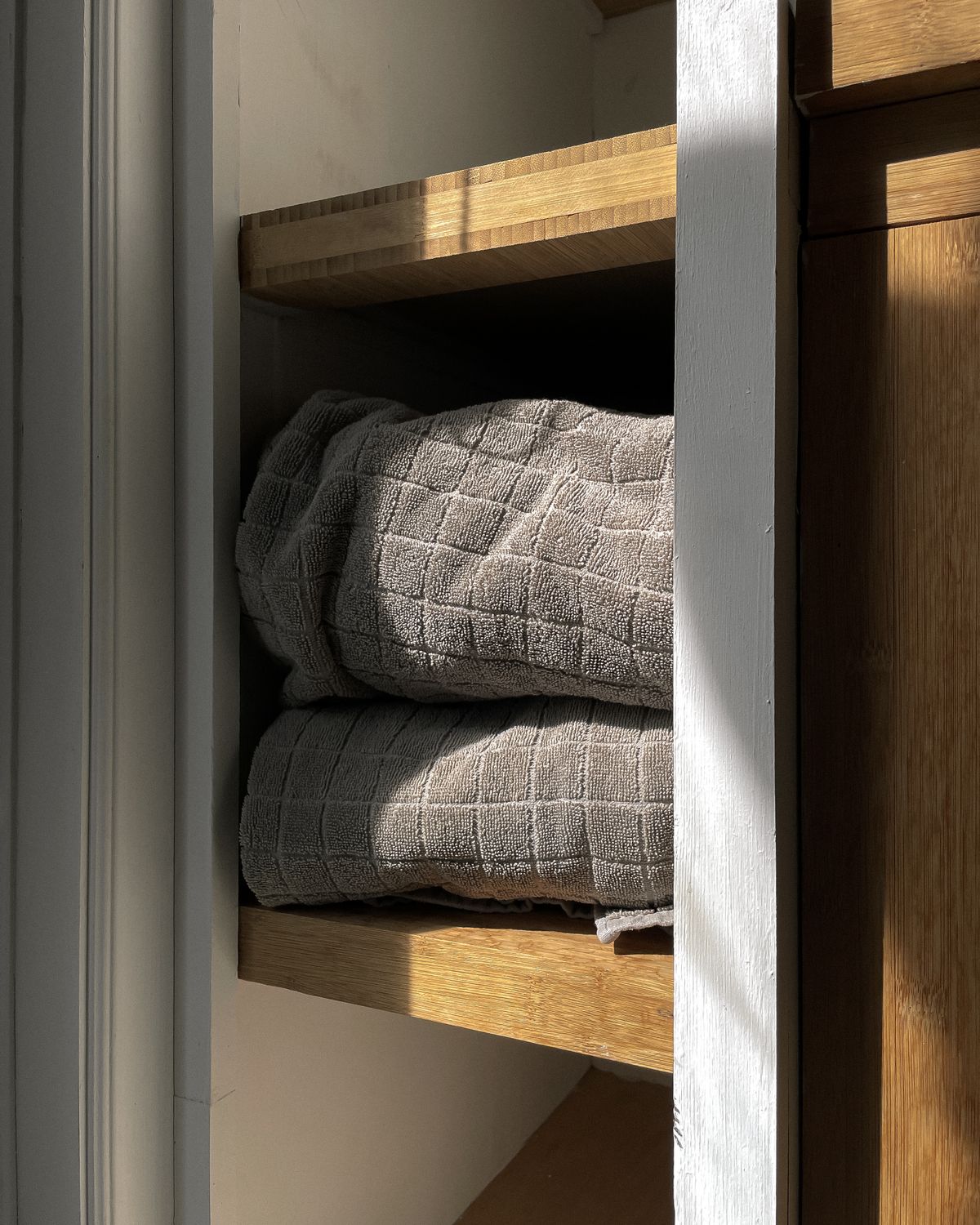 Soft gray towels neatly folded and rolled inside a sunlit wooden bathroom cabinet