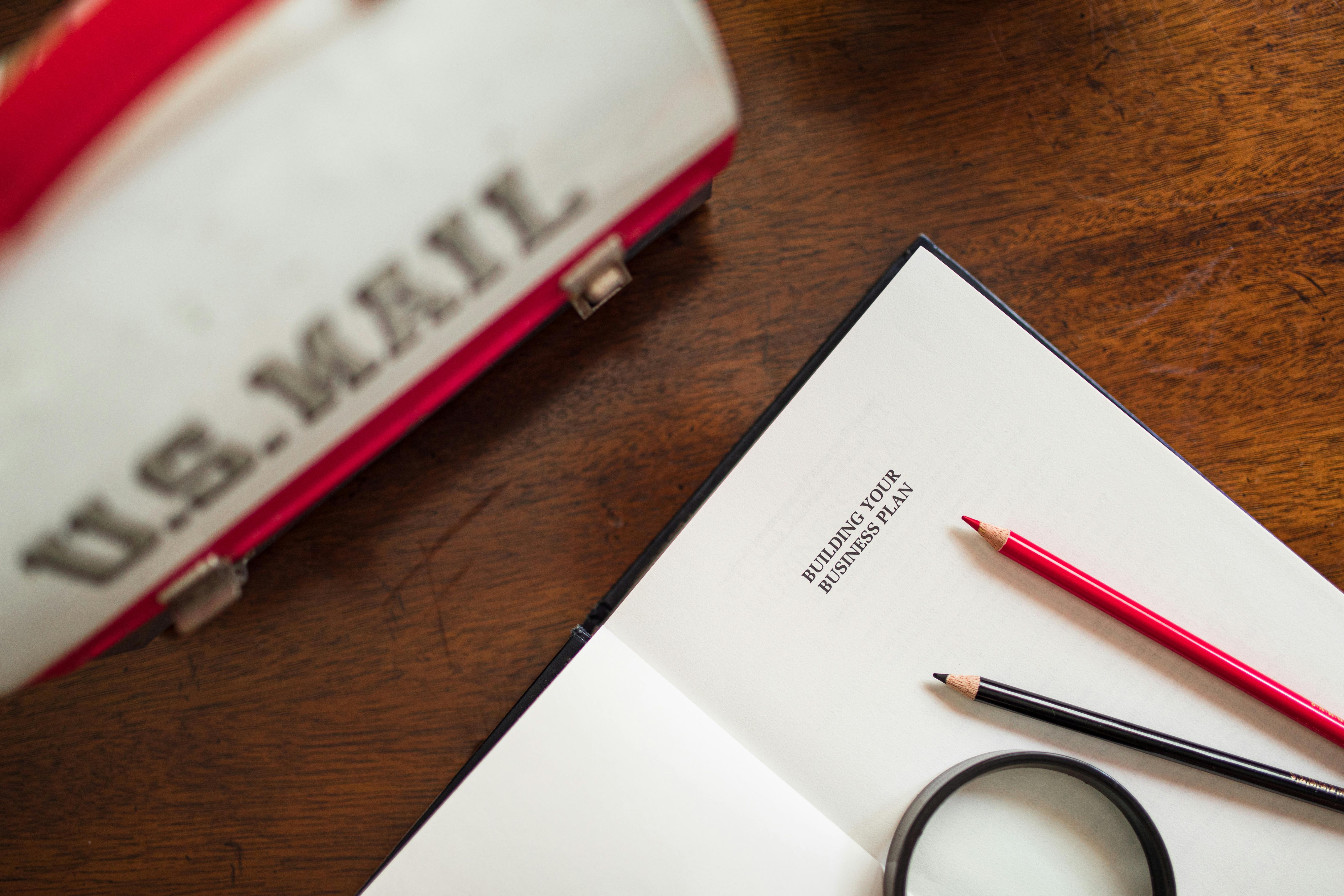 Open book with 'Building Your Business Plan' text, pencils, and U.S. Mail bag on a wooden desk.