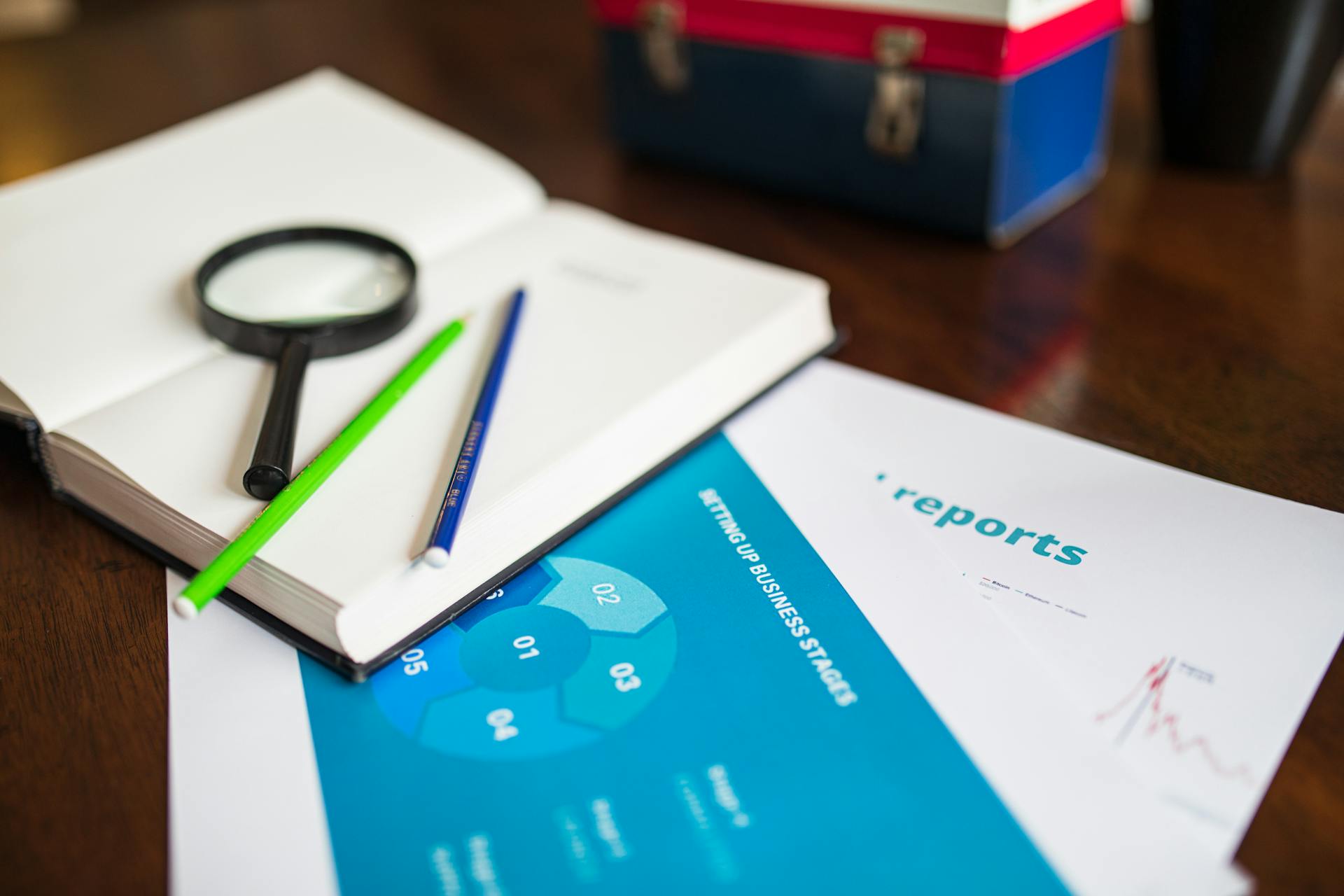 Open book with magnifying glass, pencils, and a business report on a wooden desk.