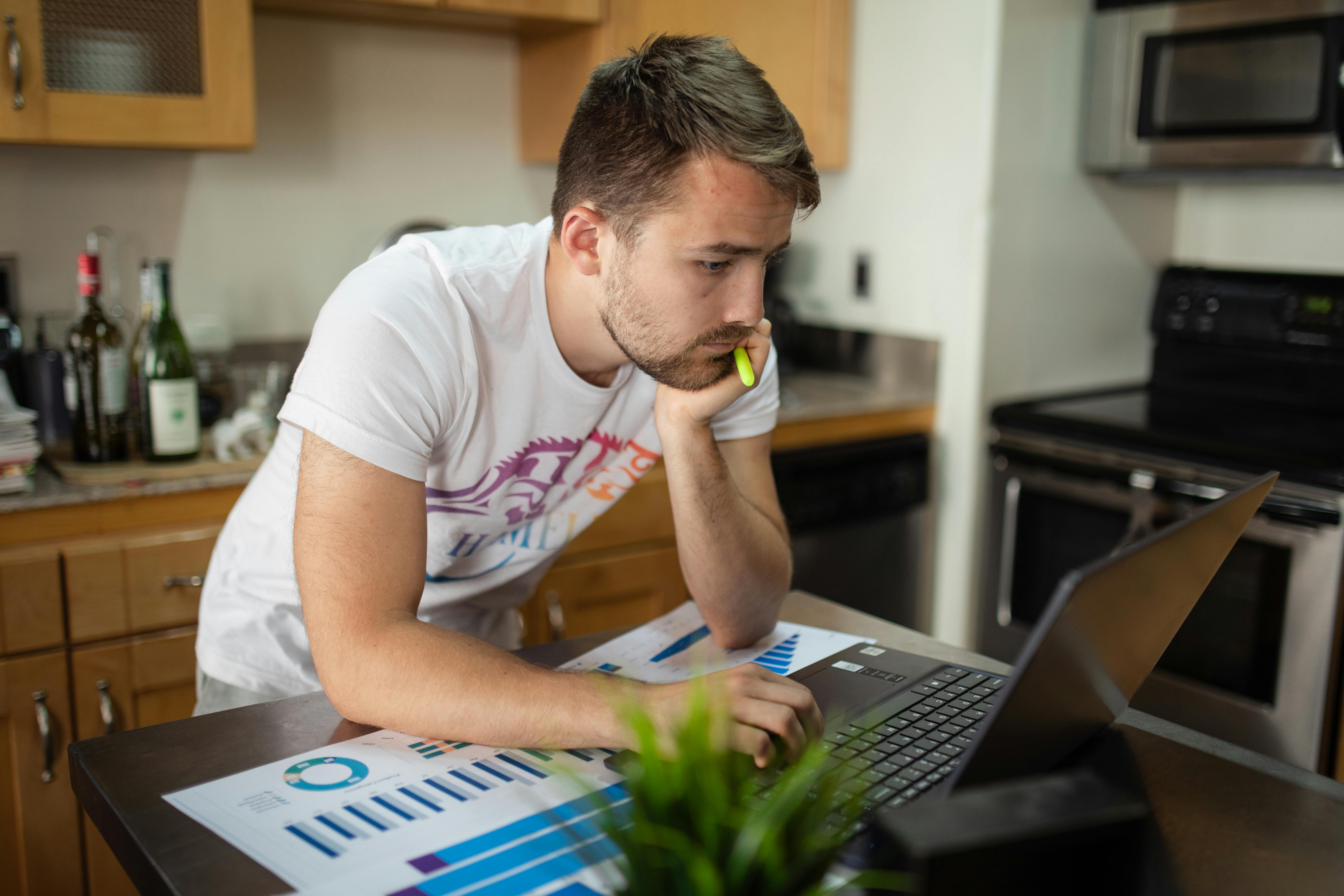 man in a white shirt looking at his laptop while his hand is on his chin