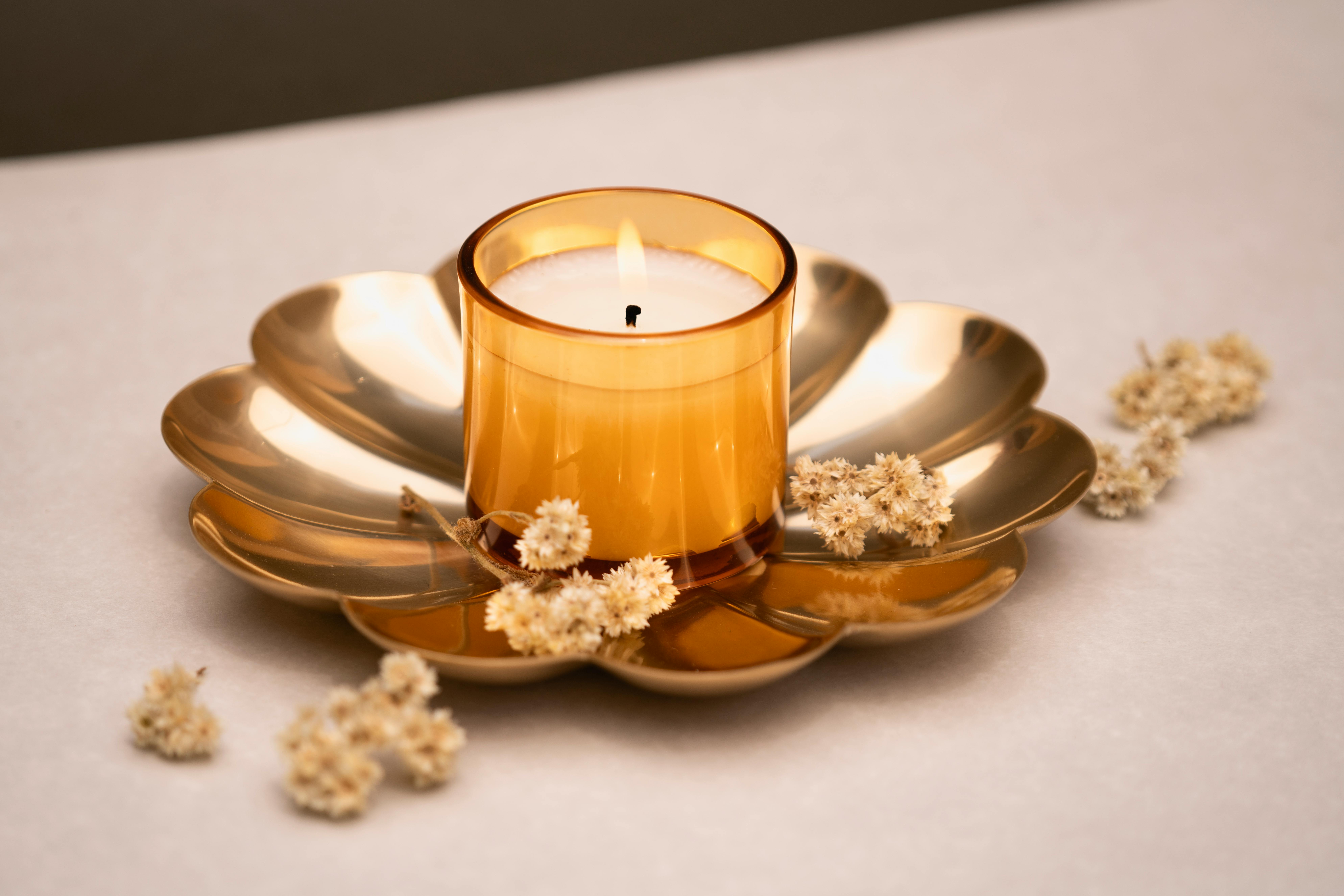 A lit candle in a golden holder surrounded by dried flowers on a white surface.
