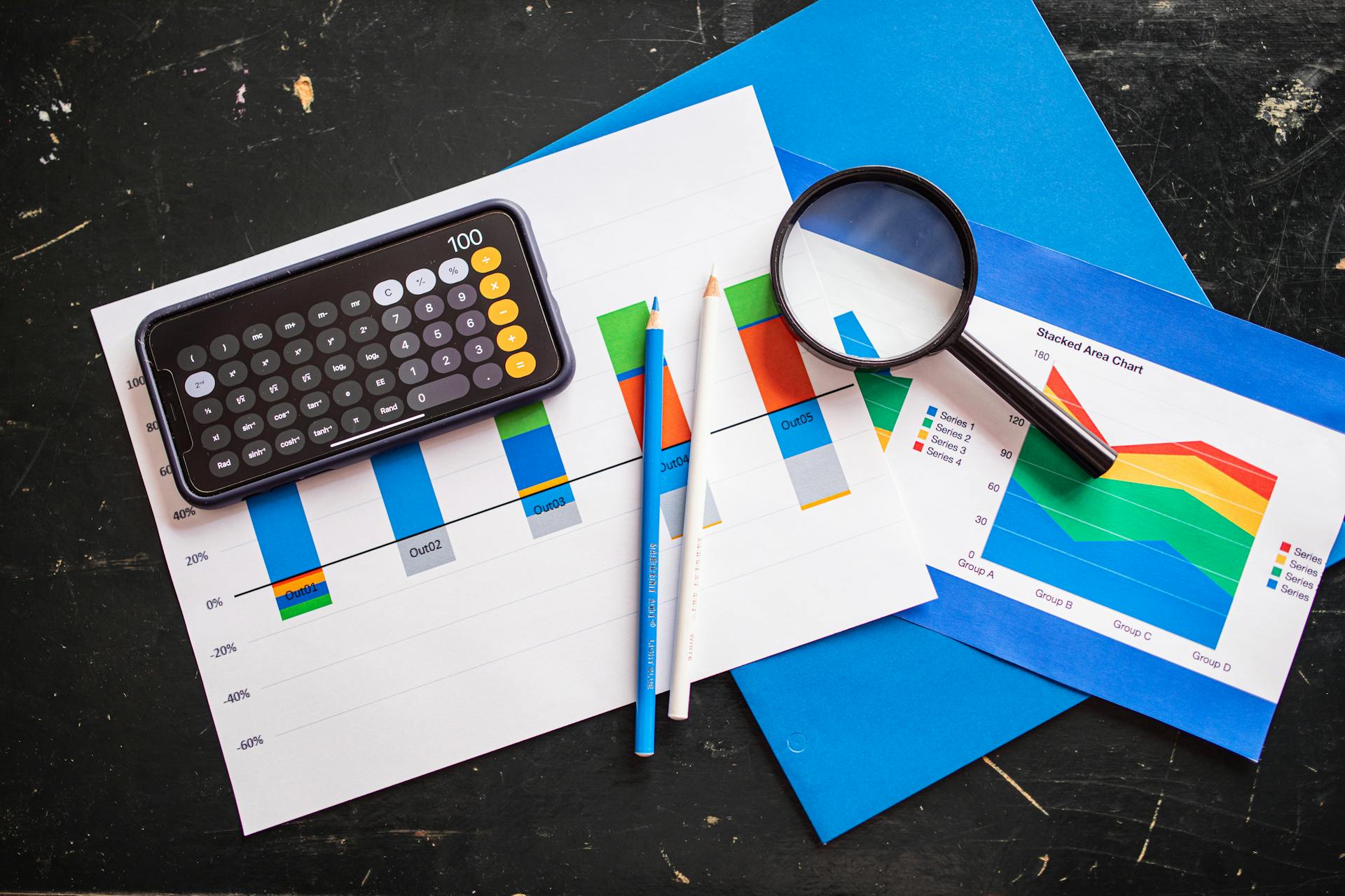 Top view of financial charts with a smartphone calculator, magnifying glass, and pencils on a desk.