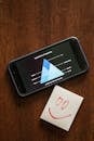 Black Android Smartphone on Brown Wooden Surface Beside a Paper with Smiley