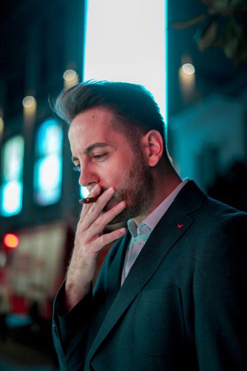 Free Pensive guy in classic jacket looking down while smoking on blurred street in evening Stock Photo