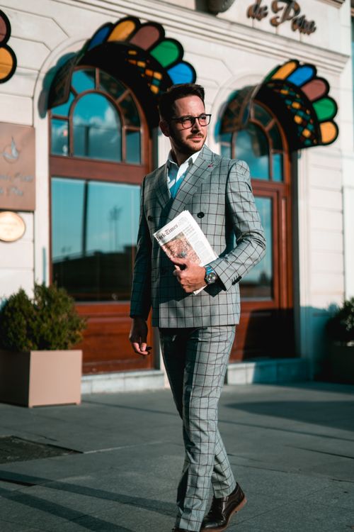 Free A fashionable man in a plaid suit holding a newspaper, walking past a colorful storefront. Stock Photo