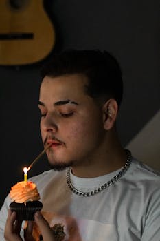 Portrait of a young man lighting a candle on a cupcake indoors, captured with a moody atmosphere.
