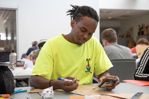 Free Focused young man sketching and studying, using a smartphone at a busy indoor workspace. Stock Photo