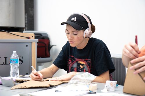 Free Young woman in headphones crafting at a table with cardboard for a school project, indoors. Stock Photo