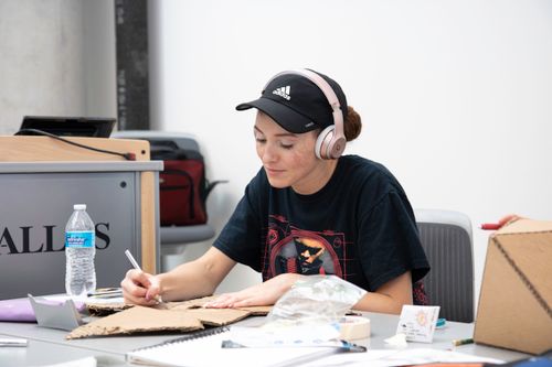 Free A young woman wearing headphones writes on cardboard at a desk, concentrating on her task. Stock Photo