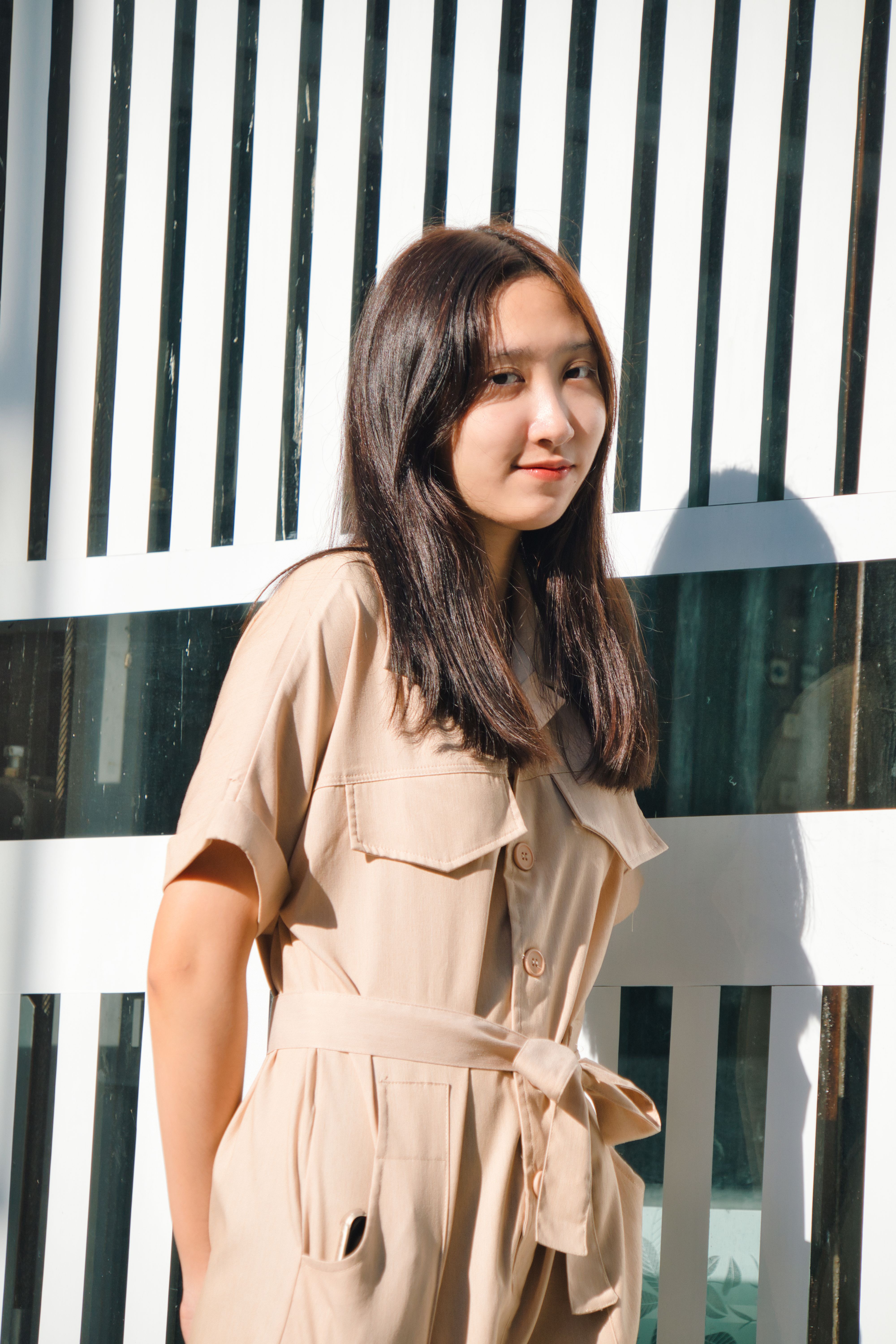 Portrait of a fashionable young woman standing against a patterned background outdoors in sunlight.